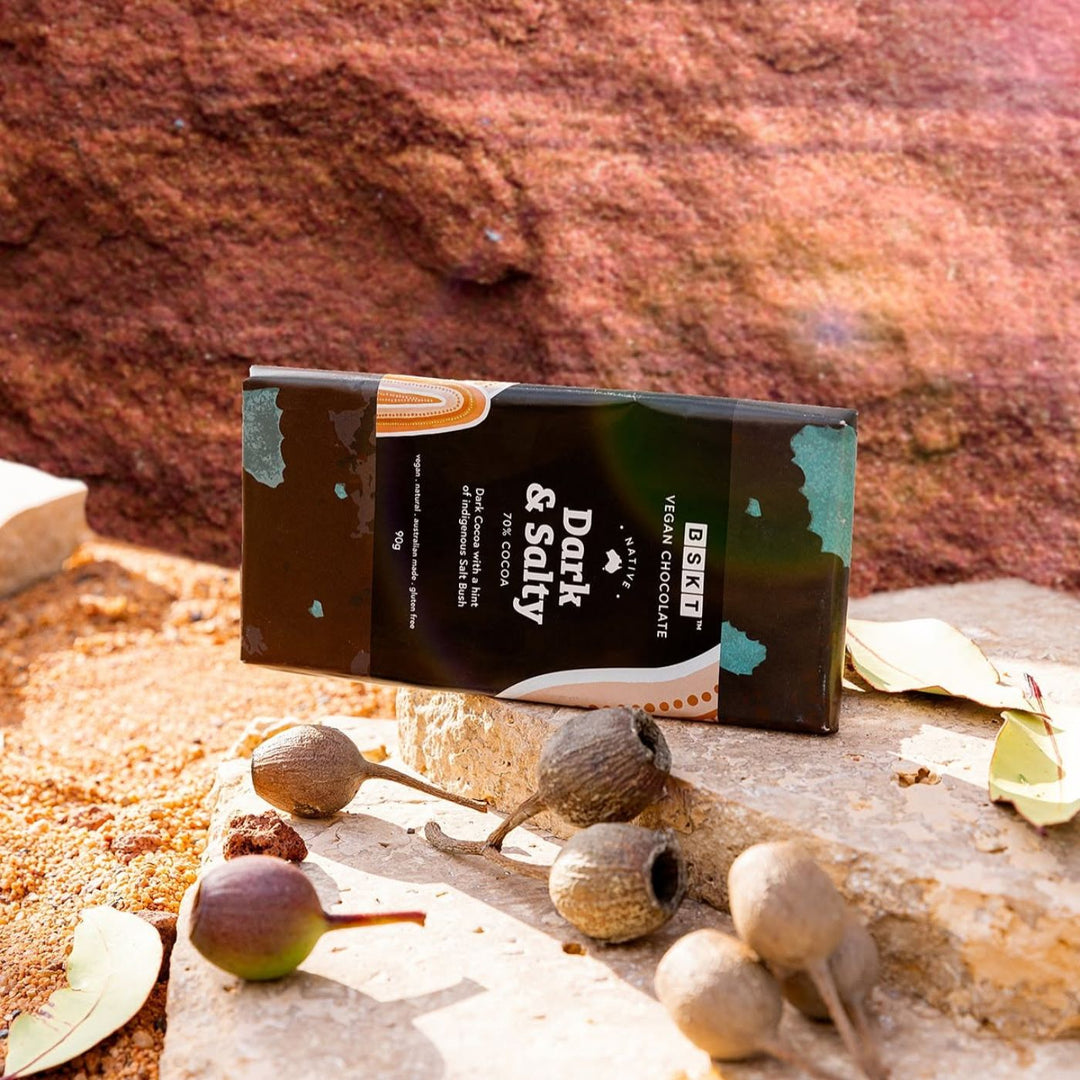 BSKT Native Dark & Salty vegan chocolate bar, 70% cocoa, standing upright against red rock, surrounded by native gum nuts and eucalyptus leaves in warm outdoor light.
