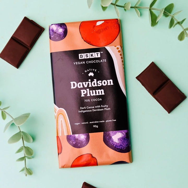 BSKT Native Davidson Plum vegan chocolate bar, 70% cocoa, in orange and purple fruit-print packaging, lying on a mint green surface surrounded by eucalyptus leaves and broken pieces of dark chocolate.