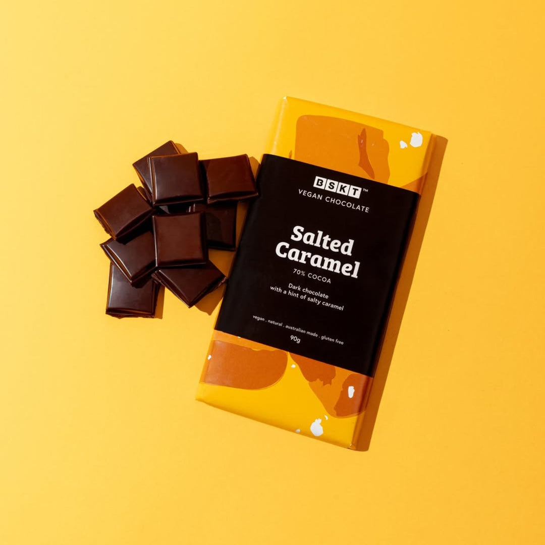 BSKT Salted Caramel vegan chocolate bar, 70% cocoa, in yellow and orange abstract-print packaging, lying on a yellow surface next to a pile of broken dark chocolate squares.