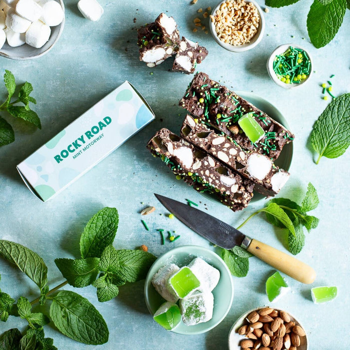 A display of Pebbly Path Mint Motorway Rocky Road bars surrounded by fresh mint leaves, green jelly pieces, almonds, and sprinkles. The rocky road bars show visible marshmallows, nuts, and green sprinkles. Product boxes are placed beside the treats. The scene is styled with green tones and natural textures.