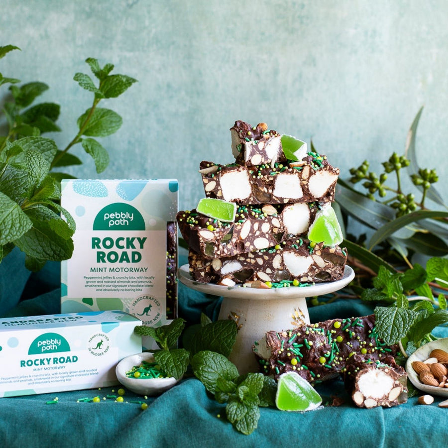 A display of Pebbly Path Mint Motorway Rocky Road bars arranged on a small cake stand and tabletop, surrounded by fresh mint leaves, green jelly pieces, almonds, and sprinkles. The rocky road bars show visible marshmallows, nuts, and green sprinkles. Product boxes are placed beside the treats. The scene is styled with green tones and natural textures.