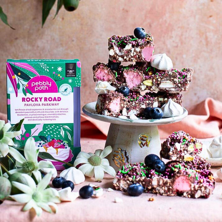 A display of Pavlova Parkway rocky road pieces stacked on a small cake stand, surrounded by blueberries, mini meringues and sprinkles. The rocky road shows visible chunks of marshmallow, nuts, meringue and dried fruit coated in chocolate. The Pebbly Path Pavlova Parkway product box stands beside the display. The scene includes flowers and soft pink fabric in the background.