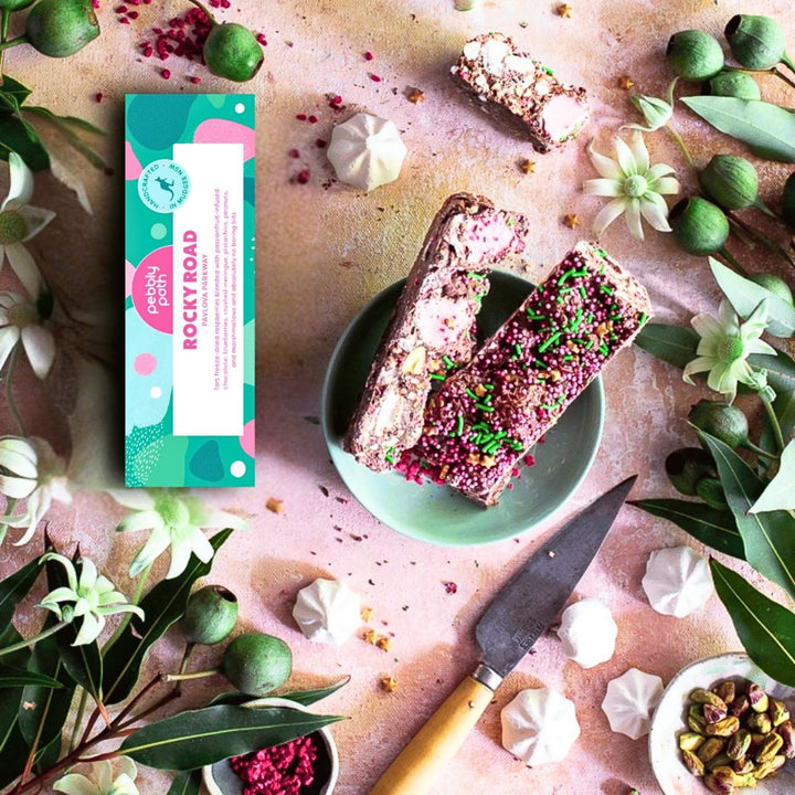 A display of Pavlova Parkway rocky road pieces surrounded by blueberries, mini meringues and sprinkles. The rocky road shows visible chunks of marshmallow, nuts, meringue and dried fruit coated in chocolate. The scene includes flowers and soft pink fabric in the background.