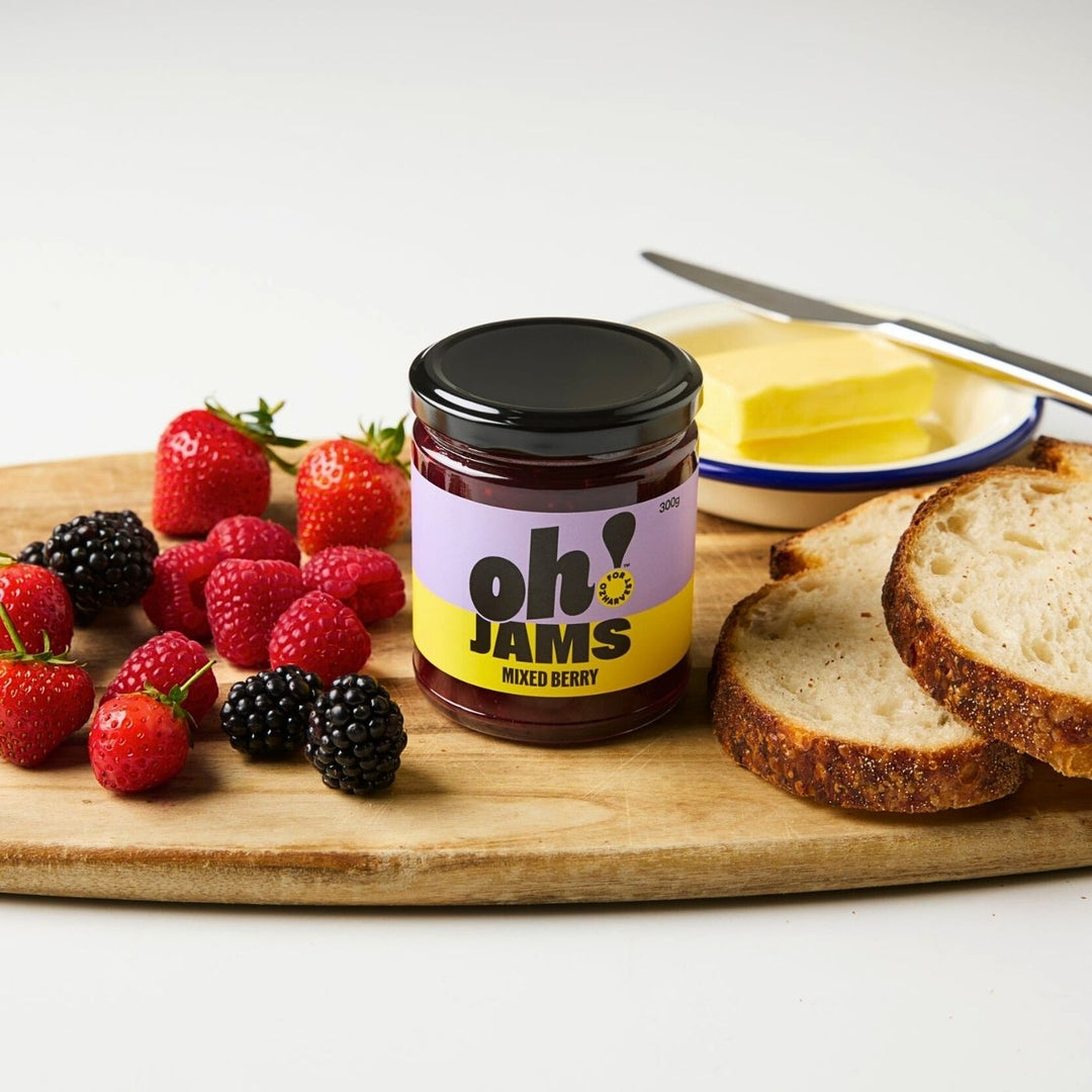 Jar of Oh! Jams Mixed Berry jam on a wooden board with strawberries, raspberries, blackberries, sliced bread and a plate of butter.