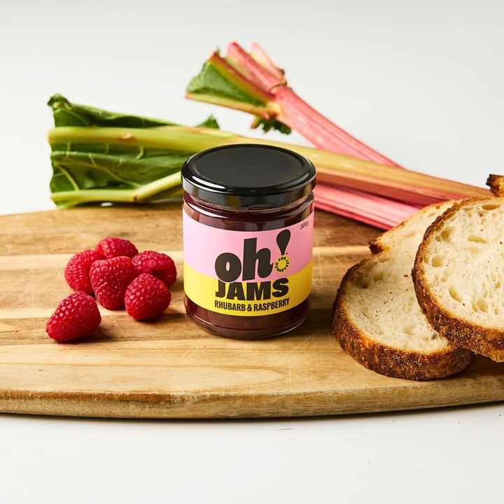 Jar of Oh! Jams Rhubarb and Raspberry jam on a wooden board with fresh raspberries, rhubarb stalks and sliced bread.”