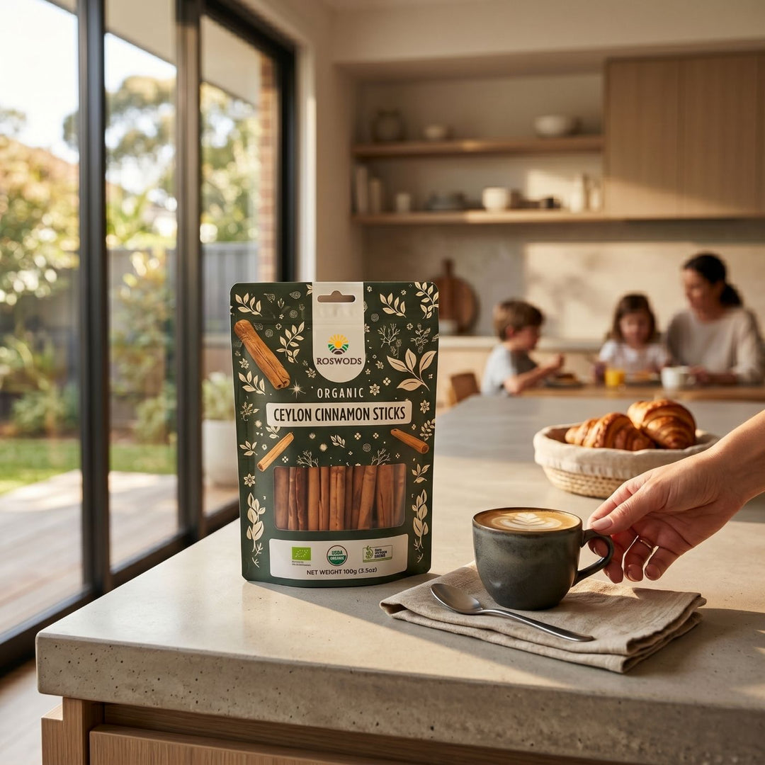Roswods Organic Ceylon Cinnamon Sticks 100g pack on a kitchen counter beside a latte being picked up, with croissants and a family at a breakfast table in the background of a bright, modern Australian home.