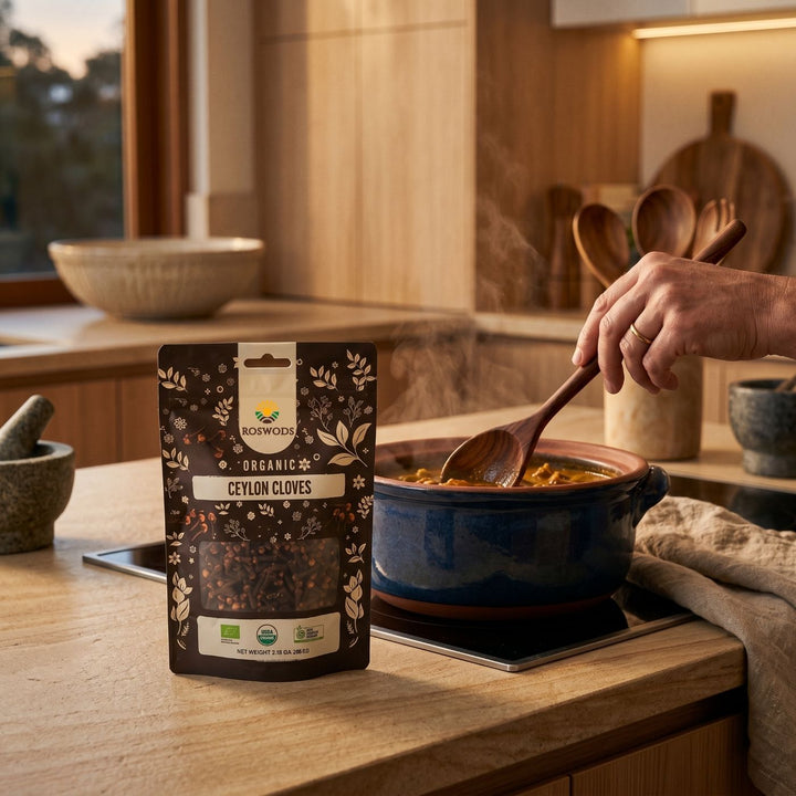 Roswods Organic Ceylon Cloves pack on a kitchen counter beside a steaming pot of curry being stirred with a wooden spoon, with a mortar and pestle and wooden utensils in the background of a warm, dimly lit kitchen.