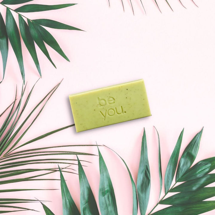 Unlabelled exfoliating body bar with lemongrass and lemon myrtle on pink background with tropical leaves, natural exfoliating soap for glowing skin